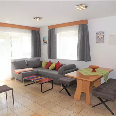 Ein helles Wohnzimmer mit einem grauen Sofa und bunten Kissen. Ein Tisch mit Stühlen ergänzt den gemütlichen Raum.