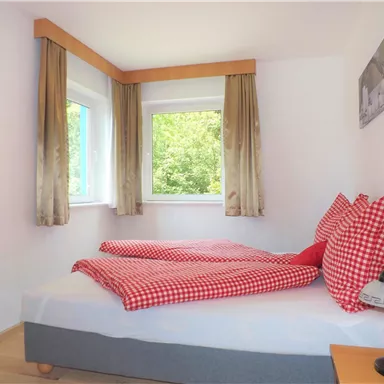 Ein helles Schlafzimmer mit zwei Einzelbetten und gestreiften roten Kissen. Die großen Fenster bieten einen Blick ins Grüne.