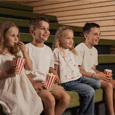 Eine Gruppe von vier Kindern sitzt auf einer Treppe und genießt Popcorn. Sie schauen gespannt in eine Richtung und scheinen eine gute Zeit zu haben.