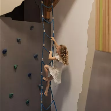 Ein Kind klettert an einer blauen Leiter in einem modernen Raum. Im Hintergrund sind Klettersteine und farbige Wände zu sehen.