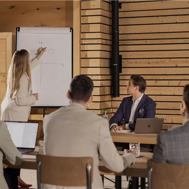 Eine Frau präsentiert vor einer Gruppe von Männern in einem modernen Konferenzraum. Sie steht neben einem Flipchart und zeigt eine Zeichnung.