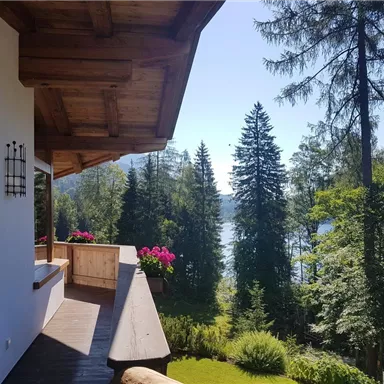 A beautiful view from a terrace with flowers and green trees. In the background, a clear lake and a blue sky can be seen.
