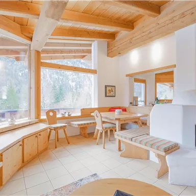 An inviting living and dining room with large windows and a wooden beam ceiling. The cozy furnishings and the view of nature create a warm atmosphere.