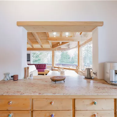 A bright kitchen with wooden decor and a spacious counter. In the background, there are cozy seating options and large windows overlooking nature.