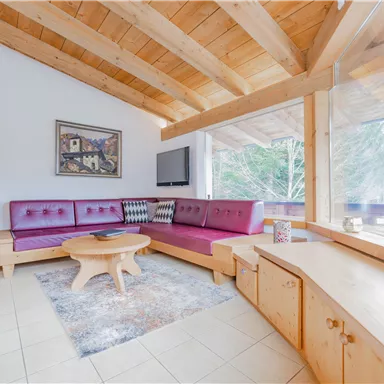 A cozy living room with wooden ceilings and modern furniture. Large windows provide a view of nature outside.