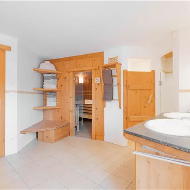 A modern bathroom with wooden paneling and two sinks. There are shelves with towels and access to another room.