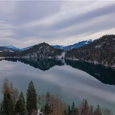 A calm lake surrounded by snow-capped mountains and pine trees. The sky is cloudy and the water reflects the landscape.