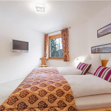 A cozy bedroom with two beds and colorful cushions. The room has a window overlooking the trees and a modern TV set on the wall.