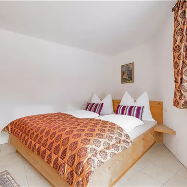A cozy bedroom with a double bed and colorful pillows. The walls are simple, and there is a large window with curtains.