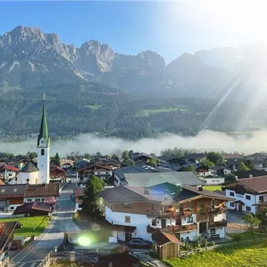 A picturesque mountain landscape with a small village in the foreground. The sun is shining over the rooftops and the church with its green tower.