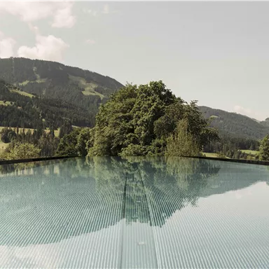 Ein Infinity-Pool mit klarem Wasser spiegelt die umliegende Landschaft. Im Hintergrund sind sanfte Hügel und grüne Bäume zu sehen.