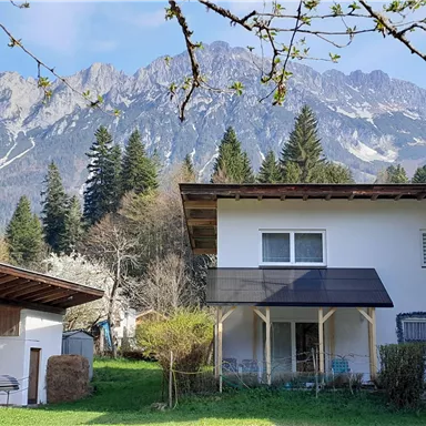 Eine ruhige Wohngegend mit einem weißen Haus und einem grünen Garten. Im Hintergrund erheben sich majestätische Berge unter einem klaren Himmel.