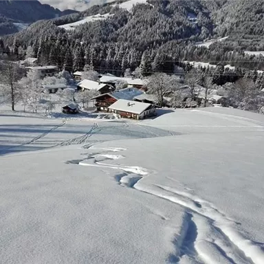 Eine winterliche Landschaft mit schneebedeckten Hügeln und einem kleinen Bauernhof im Hintergrund. Die Umgebung ist ruhig und malerisch, umgeben von schneebedeckten Bäumen.