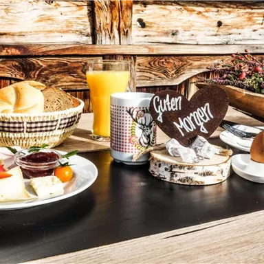 Ein reichhaltiges Frühstück mit Brötchen, Eiern und frischem Orangensaft. Auf dem Tisch steht ein Holzschild mit der Aufschrift "Guten Morgen".