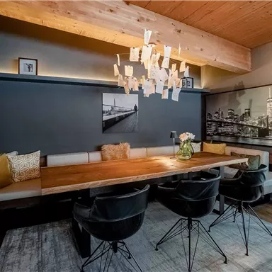 Ein modernes Esszimmer mit einem großen Holztisch und schwarzen Stühlen. Die Wände sind in einem dunklen Farbton gehalten, und eine auffällige chandelier sorgt für Beleuchtung.