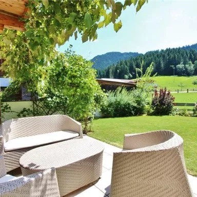 Ein einladender Garten mit gemütlichen Sitzmöbeln aus Rattan. Im Hintergrund sind grüne Wiesen und bewaldete Hügel zu sehen.