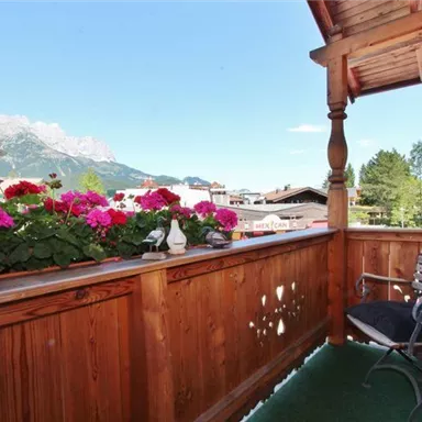 Ein gemütlicher Balkon mit blühenden Pflanzen und einem Blick auf die Berge. Die Holzverkleidung und der Stuhl laden zum Entspannen ein.