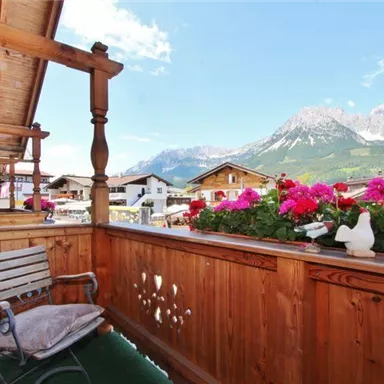 Ein schöner Balkon mit Holzgeländer und blühenden Blumen. Im Hintergrund sind majestätische Berge und ein klarer Himmel zu sehen.