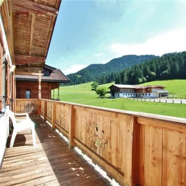 Ein sonniger Balkon mit Holzgeländer und Blick auf grüne Wiesen und Berge. Im Hintergrund sind typische alpine Häuser zu sehen.