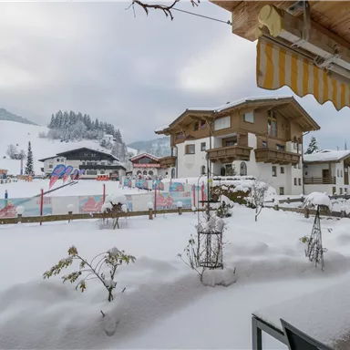 Eine winterliche Landschaft mit viel Schnee und Blick auf traditionelle Alpine Häuser. Im Hintergrund sind schneebedeckte Berge und eine Skianlage zu sehen.