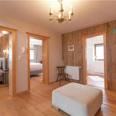 Ein gemütlicher Raum mit Holzwänden und einem modernen Kronleuchter. Im Hintergrund sind zwei Schlafzimmer und eine Sitzgelegenheit zu sehen.