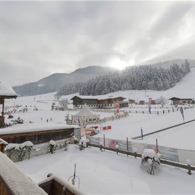 Eine winterliche Landschaft mit viel Schnee und schneebedeckten Hügeln. Im Hintergrund sind Hütten und Skispuren sichtbar, während die Sonne durch die Wolken scheint.