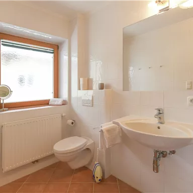 Ein modernes Badezimmer mit weißen Fliesen und einem großen Fenster. Es gibt ein Waschbecken, eine Toilette und eine Heizung.