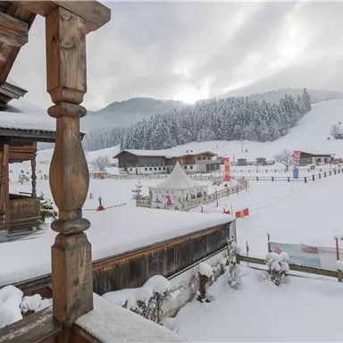 Eine verschneite Winterlandschaft mit Bergkulisse und traditionellen Holzhäusern. Im Vordergrund ist ein Skigebiet mit einem Zelt und vielen schneebedeckten Bäumen zu sehen.