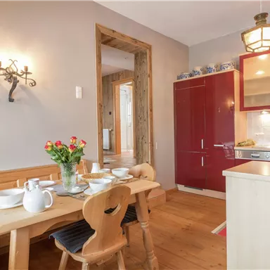 Eine gemütliche Küche mit einem Holztisch und Stühlen. Die Wände sind in sanften Farben gehalten, und es gibt einen roten Kühlschrank sowie frische Blumen auf dem Tisch.