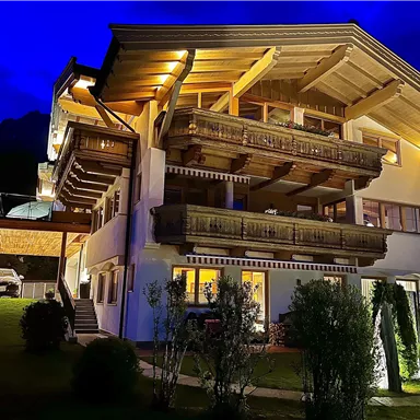 Ein wunderschönes Chalet mit Holzverkleidung und Balkonen, beleuchtet bei Nacht. Der Garten ist gepflegt und bietet eine einladende Atmosphäre.