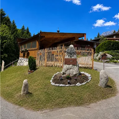 A rustic wooden house surrounded by trees and a well-maintained garden. In the foreground, there is a decorative stone with flowers and shrubs.