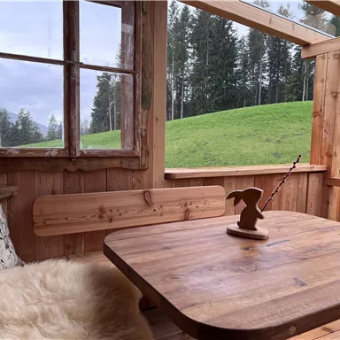 A cozy wooden room with a table and a bench. Through the window, one looks out onto a green meadow and trees.