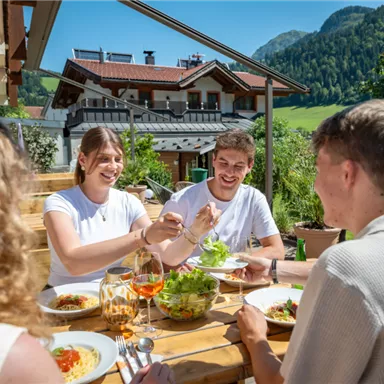 A group of friends is enjoying a meal together on a terrace with a beautiful view. The table is set with delicious dishes and drinks.