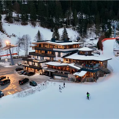 Ein modernes Berghotel in verschneiter Landschaft mit vielen Tannen. Skifahrer genießen die Piste vor dem Hotel und es stehen mehrere Autos auf dem Parkplatz.