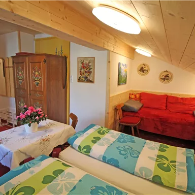 A cozy room with two beds and colorful blankets. In the room, there is a small table and a red sofa, along with wooden paneling and flowers as decoration.