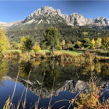A picturesque mountain landscape with tall peaks and colorful trees in the foreground. The calm lake reflects the beauty of nature.