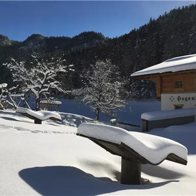 A snowy winter landscape with a cozy wooden house. In the foreground, there are two benches and a playground, surrounded by snow-covered trees.
