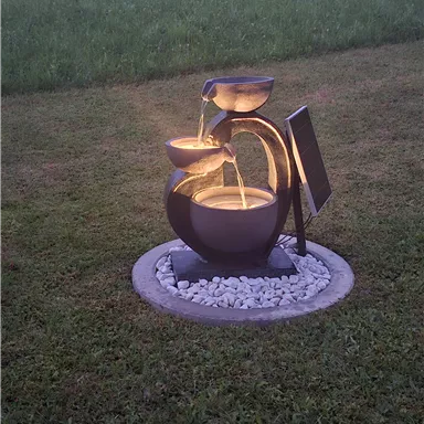 A modern garden fountain with several basins that bubble water. In the background, there is a green lawn and a wooded area.