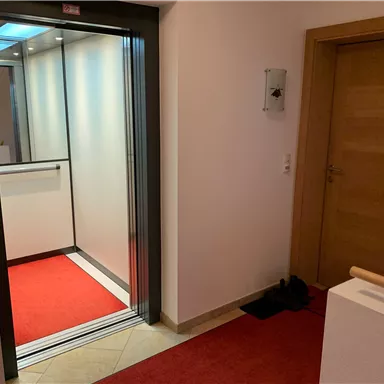 Ein moderner Aufzug in einem hellen Flur mit rotem Teppich. Auf der rechten Seite ist eine Holz- und Glastür sichtbar.