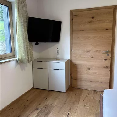 Ein schlichtes Zimmer mit einem Fernseher an der Wand und einem Holztür. Der Boden ist aus Holz und es gibt einen kleinen Schrank unter dem Fenster.
