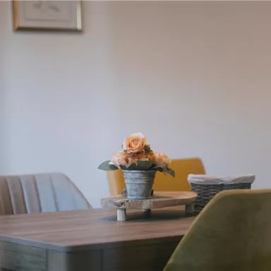 Ein schöner Tisch mit einem Blumenarrangement in einem Topf. Im Hintergrund sind Stühle und eine gedeckte Umgebung zu sehen.