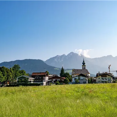 Eine malerische Landschaft mit einem Dorf im Vordergrund und majestätischen Bergen im Hintergrund. Der Himmel ist klar und die Wiese grün und einladend.