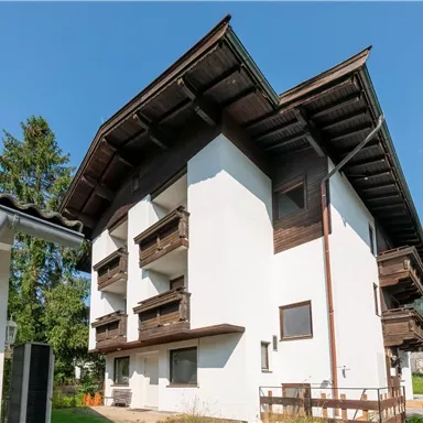 Ein modernes Gebäude mit traditionellem alpinen Stil und braunem Holz. Im Hintergrund sind Berge und ein klarer blauer Himmel zu sehen.