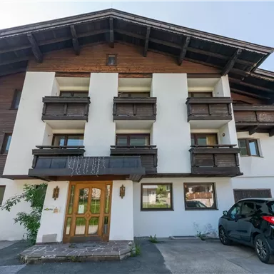 Ein modernes Gebäude im alpinen Stil mit Holzverkleidung und mehreren Balkonen. Vor dem Gebäude steht ein schwarzes Auto.