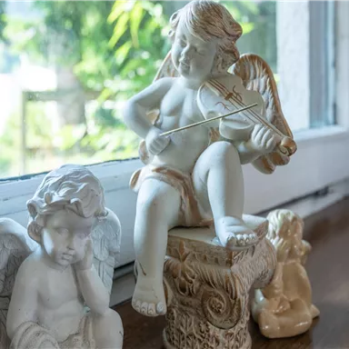 Eine Sammlung von Engeln aus Keramik, darunter ein spielender Engel mit einer Geige. Die Figuren stehen auf einer Fensterbank in einem hellen Raum.