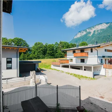 Eine Ferienhaussiedlung mit modernem Design, umgeben von grünen Wiesen und Bergen. Der Himmel ist klar und sonnig.