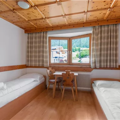 Ein einfaches Zimmer mit zwei Einzelbetten und einem kleinen Tisch. Die Fenster bieten einen Blick auf die Umgebung und lassen viel Tageslicht herein.