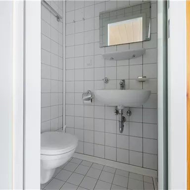 Ein kleines Badezimmer mit weißen Fliesen und einem Waschbecken. Es enthält eine Toilette und einen Spiegel über dem Waschbecken.