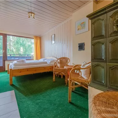 Ein gemütliches Zimmer mit einem Bett, einem Rattanstuhl und einem traditionellen Ofen. Die grüne Teppichfläche und großen Fenster schaffen eine helle, einladende Atmosphäre.