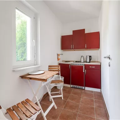 Eine kleine, moderne Küche mit roten Schränken und einem Tisch. Die Fenster bringen viel Licht in den Raum.